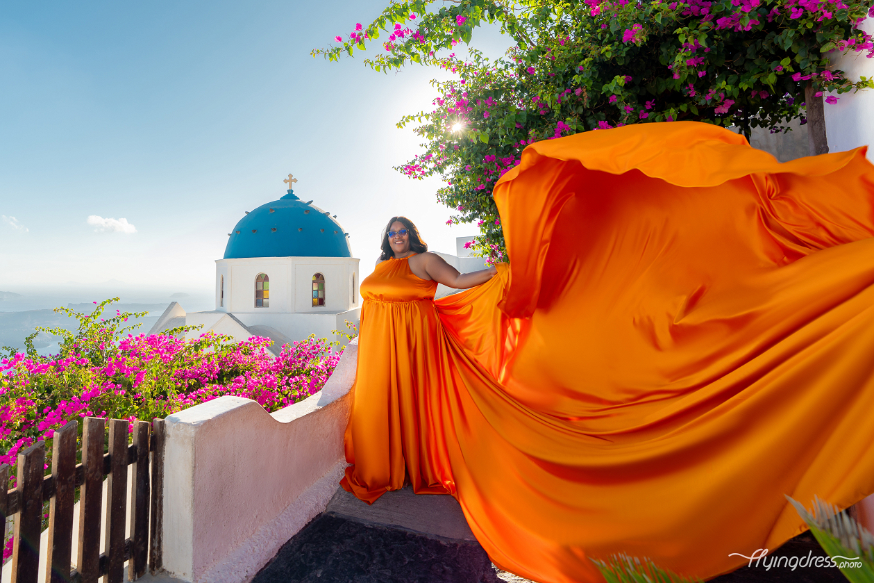 A vivid orange flying dress billows magnificently beside Santorini's iconic blue-domed church, framed by cascading fuchsia bougainvillea and golden sunlight.